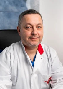 dr_mąkowski_maciej_angiolog_kraków Lekarz w białym fartuchu medycznym z logo UniCardia, siedzący w gabinecie lekarskim na tle jasnej ściany. Zdjęcie przedstawia profesjonalnego pracownika służby zdrowia podczas pracy w klinice medycznej.