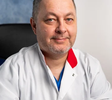 Lekarz w białym fartuchu medycznym z logo UniCardia, siedzący w gabinecie lekarskim na tle jasnej ściany. Zdjęcie przedstawia profesjonalnego pracownika służby zdrowia podczas pracy w klinice medycznej.